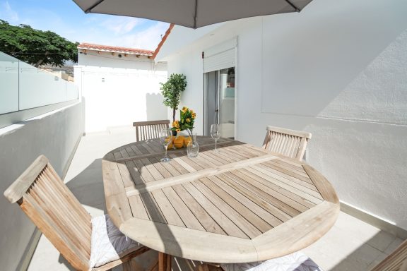 Mesa de madera con sillas bajo una sombrilla en una terraza luminosa.