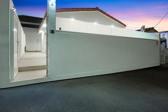 Entrada de un patio iluminado al atardecer con muro blanco y casa al fondo.