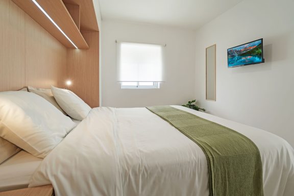 Habitación luminosa con cama, sábanas blancas y una manta verde. Televisor en la pared.