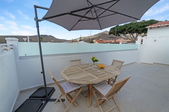Mesa de madera con sillas bajo una sombrilla en una terraza.