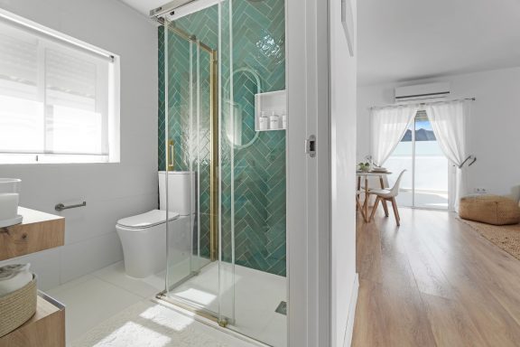 Baño moderno con ducha de azulejos verdes y salón luminoso al fondo.