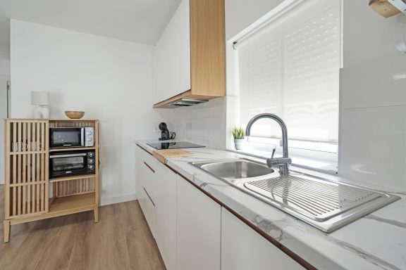 Cocina moderna con muebles claros, fregadero y estantería de madera con electrodomésticos.