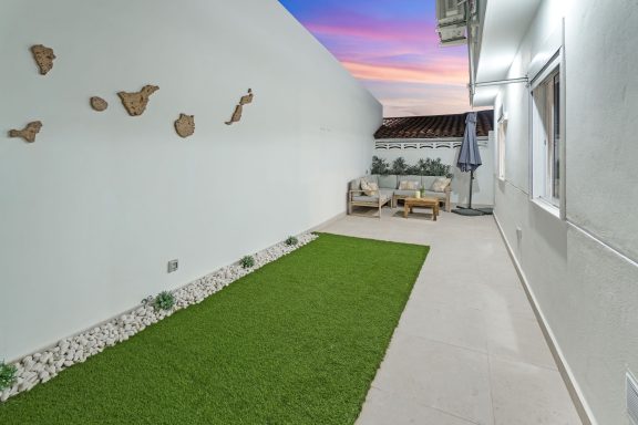 Patio con césped artificial, paredes blancas y bancos, al atardecer.