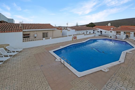 Piscina al aire libre rodeada de tumbonas y casas blancas bajo un cielo despejado.