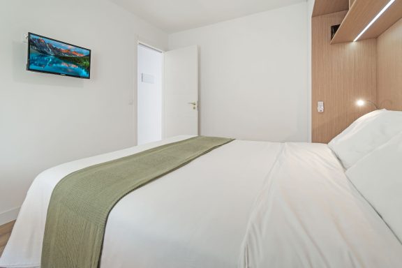 Habitación moderna con cama grande, decoración clara y televisor en la pared.