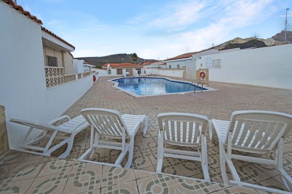 Zona de piscina con tumbonas blancas y casas alrededor, bajo un cielo despejado.