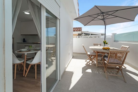 Terraza con mesa, sillas de madera y sombrilla, con vista clara y entorno luminoso.