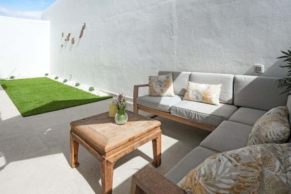 Salón exterior con sofá gris, mesa de madera y césped artificial en un ambiente luminoso.